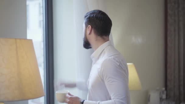 Un hombre de negocios seguro y guapo bebiendo café de la mañana junto a la ventana de la habitación del hotel. Éxito joven caucásico hombre en camisa blanca elegante en viaje de negocios. Viajar, estilo de vida, comodidad, negocios . — Vídeos de Stock