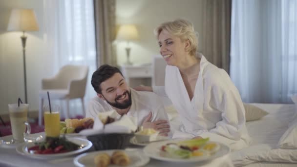 Šťastná kavkazská nevěsta a ženich si užívají líbánky v luxusním hotelu. Usmívající se krásná žena a pohledný muž klábosení a jíst čerstvé ovoce ráno. Pokojová služba, štěstí, svatba. — Stock video