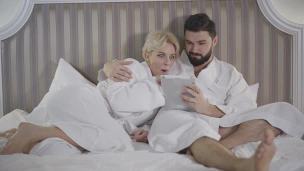 Spannende blanke vrouw toont iets op tabletscherm aan liefhebbende man. Mooi glimlachend echtpaar rustend in bed in hotelkamer of thuis en pratend. Vreugde, levensstijl, vrije tijd, reizen, toerisme. — Stockvideo