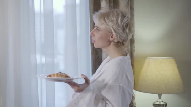 Vista lateral da linda mulher loira olhando pela janela e comendo croissant delicioso fresco. Jovem senhora caucasiana desfrutando de café da manhã no hotel no resort. Estilo de vida, felicidade, lazer, relaxamento . — Vídeo de Stock