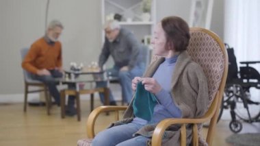 Arka planda satranç oynayan yaşlı beyaz kadın portresi örgü örüyor ve erkeklere bakıyordu. Huzurevindeki emeklilerin yaşam tarzı.