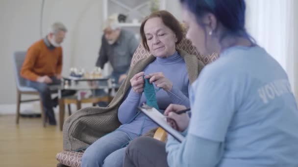 Voluntaria caucásica borrosa hablando con una jubilada en un asilo. Mujer anciana tejiendo y haciendo un gesto no agitando la cabeza . — Vídeos de Stock