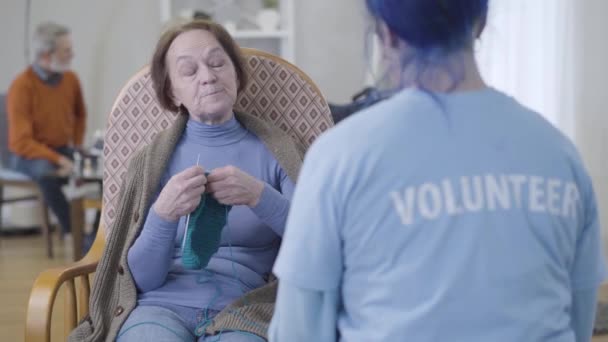 Retrato de una mujer caucásica madura positiva hablando con un voluntario irreconocible en un asilo de ancianos. Jubilado mayor tejer y charlar emocionalmente . — Vídeos de Stock