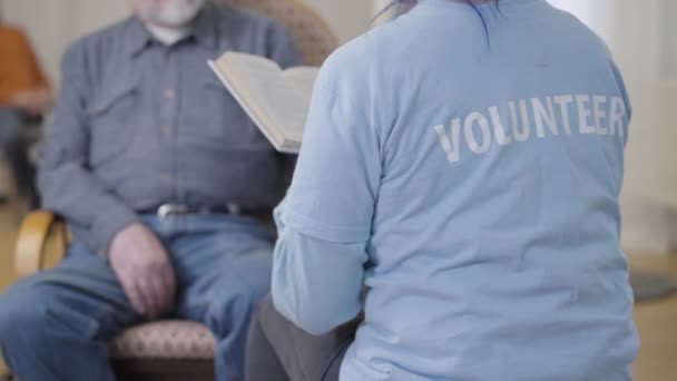 Tanımlanamayan gönüllü, koltukta oturan bulanık yaşlı adam için kitap okuyor. Huzurevinde gönüllü olarak çalışmak. Edebiyat okuyucusunu dinleyen olgun bir emekli.. — Stok video