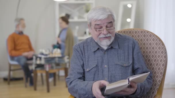 Portret dojrzałego białego mężczyzny w okularach czytającego książkę w domu opieki. Starszy mężczyzna emeryt chętnie literatury korzystających z wolnego czasu. — Wideo stockowe