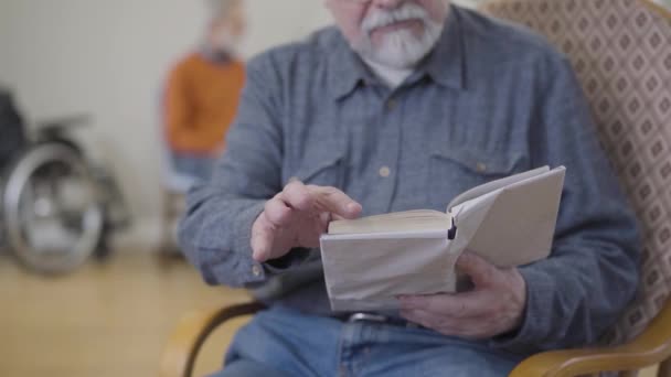 Сконцентрированный неузнаваемый старый кавказский мужчина, читающий литературу в доме престарелых. Размытый мужчина-пенсионер в очках отдыхает в доме престарелых. Окутанный книгой . — стоковое видео