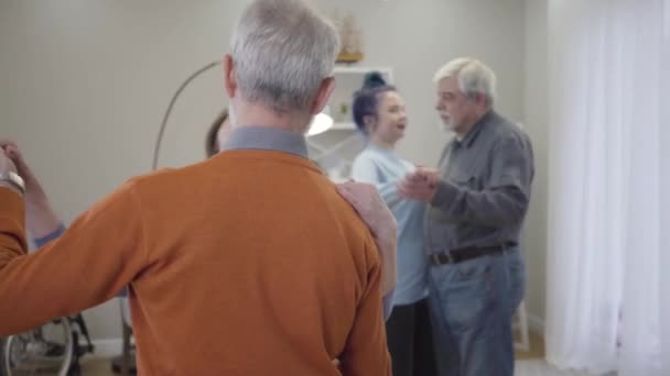 Щасливий кавказький старший чоловік і жінка танцюють у будинку престарілих. Самотні літні пенсіонерські танці з добровольцями на задньому плані. Старі пенсіонери насолоджуються вечором. — стокове відео