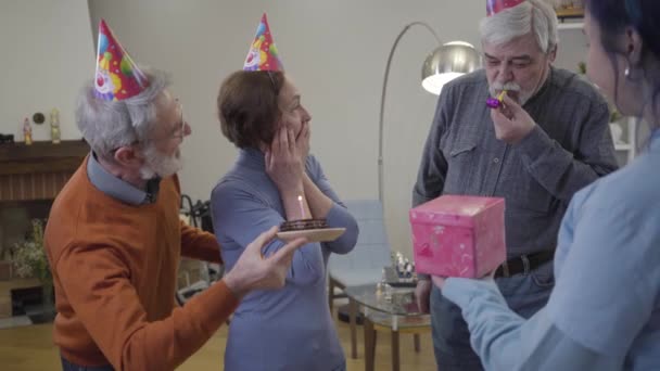 Excited happy old Caucasian woman celebrating birthday in nursing home. Residents and volunteer blowing party whistle and presenting gift and cake. — Stock Video