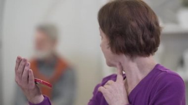 Son sınıf öğrencisi beyaz bir kadının el aynasına bakışını yakından çek. Kadın emekli, bulanık adama dönüp makyajını kontrol ediyor..