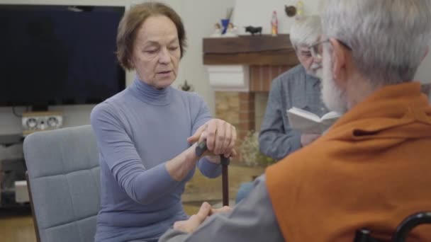 Beyaz saçlı, beyaz, olgun bir adam yaşlı kadını bastonla sakinleştiriyor. Huzurevinde birbirlerine destek olan yaşlı emekliler. Son sınıf öğrencisi arka planda kitap okuyor.. — Stok video