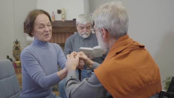 Portret van een positieve oudere blanke vrouw die praat met een volwassen man en lacht. Ouderen paar flirten als eenzame man lezen op de achtergrond in verpleeghuis. — Stockvideo