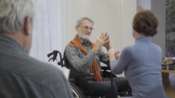 Tekerlekli sandalyedeki zarif, yaşlı beyaz adam yaşlı kadının elini tutuyor. Yetişkin bir emekli, huzurevinin bayan sakiniyle flört ediyor. Yalnız adam ön planda kitap okuyor.. — Stok video
