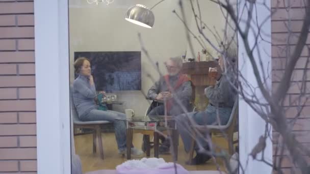 연로 한 백인 세 사람 이 요양원에서 이야기를 나누고 차를 마시고 있었다. 남자와 여자 가 실내에서 조용 한 저녁을 보내고 있습니다. 넓은 샷, 외부에서 촬영. — 비디오