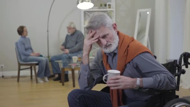 Hombre caucásico mayor agotado en silla de ruedas frotando las sienes como residentes positivos de hogares de ancianos hablando y riendo en el fondo . — Vídeo de stock