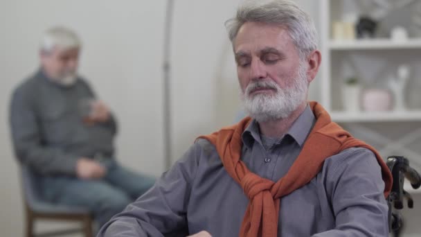 Retrato de un hombre caucásico mayor alegre mirando repentinamente a la cámara y sonriendo. Viejo positivo sentado en silla de ruedas en un asilo de ancianos con un tipo borroso al fondo . — Vídeos de Stock