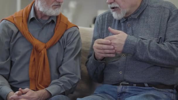 Tanımlanamayan yaşlı beyaz adam arkadaşıyla konuşuyor ve el kol hareketi yapıyor. İki emekli erkek kanepede oturup sohbet ediyorlar.. — Stok video