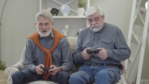 Zwei glückliche alte kaukasische Männer spielen Videospiel, geben fünf und lächeln. Positiv gelaunte Rentner haben Spaß im Haus. — Stockvideo