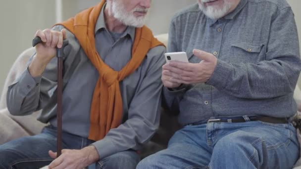 Twee onherkenbare oudere blanke mannen die smartphone gebruiken en praten. Joyful mannelijke gepensioneerden chatten op de bank in verpleeghuis. — Stockvideo