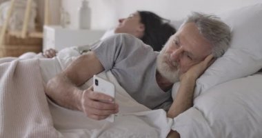 Beyaz adamın yakın plan portresi, yatakta yatan ve arka planda uyuyan karısı olarak akıllı telefonu kullanan. Modern teknolojileri kullanan yakışıklı, olgun bir adam. Boş zaman, günaydın. Sinema 4k ProRes Merkezi