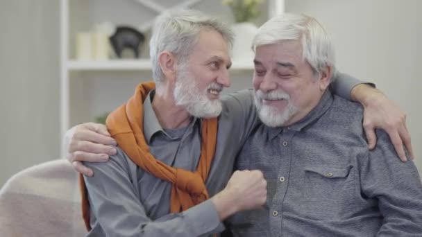 Homens brancos idosos positivos abraçando, falando e olhando para a câmera. Retrato de melhores amigos posando em casa de repouso dentro de casa . — Vídeo de Stock