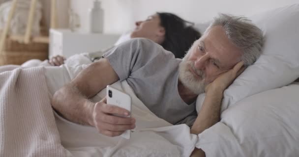 Beyaz adamın yakın plan portresi, yatakta yatan ve arka planda uyuyan karısı olarak akıllı telefonu kullanan. Modern teknolojileri kullanan yakışıklı, olgun bir adam. Boş zaman, günaydın. Sinema 4k ProRes Merkezi — Stok video