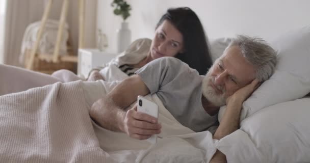 Podezřelá starší běloška se dívá přes manželovo rameno na obrazovku chytrého telefonu. Starší muž chatuje online s někým a žena nakukuje. Žárlivost, rodina, zrada. Cinema 4k ProRes HQ. — Stock video