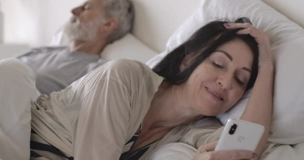 Zbliżenie portret starszej brunetki kaukaskiej kobiety używającej smartfona leżącego w łóżku jako męża śpiącego w tle. Szczęśliwa żona korzystająca z mediów społecznościowych. Styl życia, radość, poranek. Kino 4k ProRes kwatera główna. — Wideo stockowe
