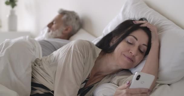 Χαμογελαστή μελαχρινή λευκή γυναίκα που χρησιμοποιεί τα μέσα κοινωνικής δικτύωσης ως ώριμο άντρα που κοιμάται. Σύζυγος ξαπλωμένη στο κρεβάτι το πρωί με smartphone και ο άνθρωπος στο παρασκήνιο. Ελεύθερος χρόνος, τρόπος ζωής. Κινηματογράφος 4k ProRes HQ. — Αρχείο Βίντεο