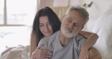 Esmer, üst düzey, beyaz, yakışıklı, gri saçlı kocasını kucaklayan bir kadın. Yetişkin çiftler sabahlarını yatak odasında geçirirler. Mutlu bir ailenin portresi. Sonsuz aşk kavramı. Sinema 4k ProRes Merkezi.