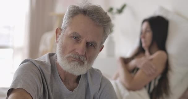 Zbliżenie przystojnego starszego siwego mężczyzny patrzącego w kamerę. Dojrzały, pewny siebie biały mężczyzna udający zamazaną żonę leżącą w łóżku w tle. Styl życia, wypoczynek. Siedziba główna Cinema4k ProRes — Wideo stockowe