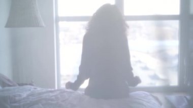 Büyük pencerenin önünde uzanan genç bir kadının silüeti. Beyaz perdenin arkasında pijamalı bir kız var. Perde açılıyor..