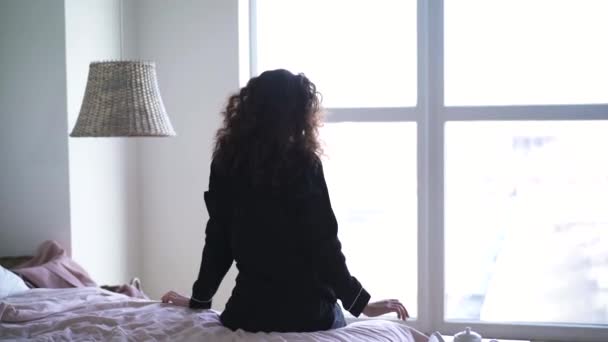 Mujer joven estirándose en la cama delante de una gran ventana. Vista trasera de la morena chica caucásica en pijama despertando en el dormitorio por la mañana . — Vídeos de Stock