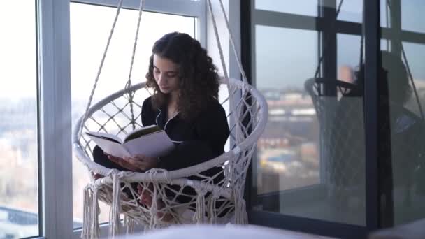 Junge kaukasische Frau sitzt im hängenden Sessel und liest Buch. Porträt eines intelligenten, literaturbegeisterten Mädchens. — Stockvideo