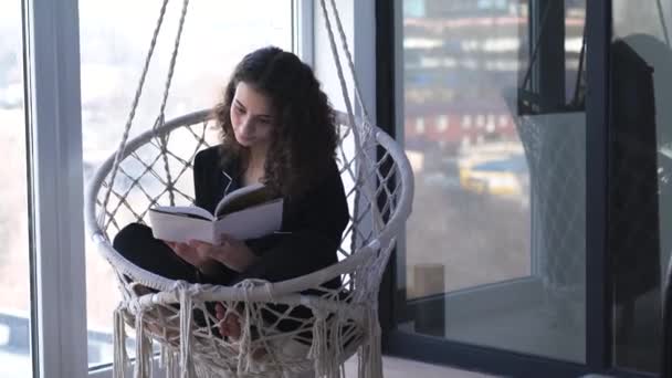 Magabiztos kaukázusi nő, aki könyvet lapoz és kinézett az ablakon. Portré fiatal gyönyörű lány olvasás lógó karosszék. — Stock videók