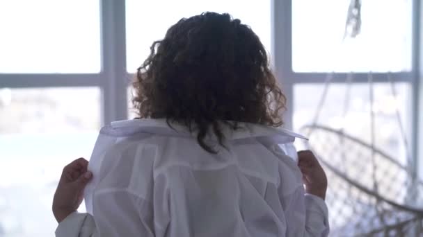 Mladá běloška si pomalu svléká bílou košili. Bruneta dívka s kudrnatými vlasy stojící před oknem ve spodním prádle. Detail, pohled zezadu. — Stock video