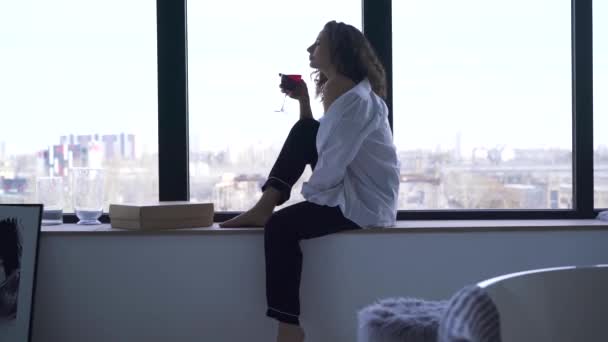 Mujer joven sentada en el alféizar de la ventana y bebiendo vino. Vista lateral de la niña morena caucásica reflexiva disfrutando de la noche tranquila en casa o en la habitación del hotel . — Vídeos de Stock