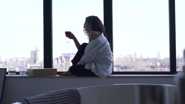 Vonzó kaukázusi lány, aki egy pohár vörösbort rázott. A fiatal barna nő élvezi a nyugodt estét az ablakpárkányon ülve. Oldalnézet. — Stock videók