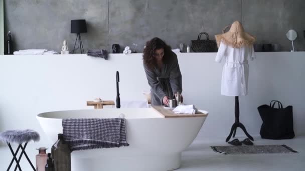 Jonge brunette vrouw in badjas het controleren van de watertemperatuur in bad. Mooi blank meisje lopend in grote badkamer. Breed schot. — Stockvideo