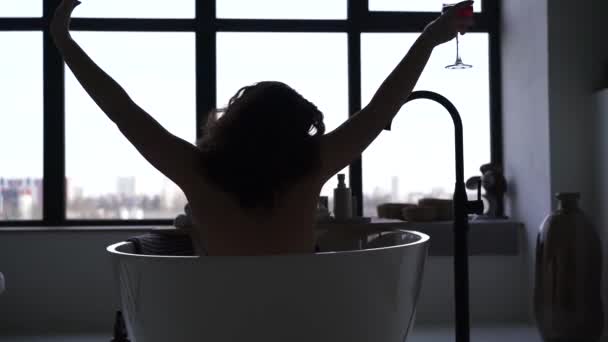 Silhouette einer jungen Frau, die Wein in der Badewanne trinkt. Fröhliches kaukasisches Mädchen, das sich abends im Badezimmer vor dem großen Fenster ausruht. Rückseite. — Stockvideo