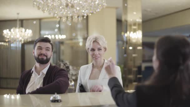 Visão traseira de recepcionista irreconhecível acolhedor clientes ricos em hotel luxuoso e dando-lhes cartão-chave do quarto. Homem e mulher felizes sorrindo e saindo como paquete passando ao fundo . — Vídeo de Stock