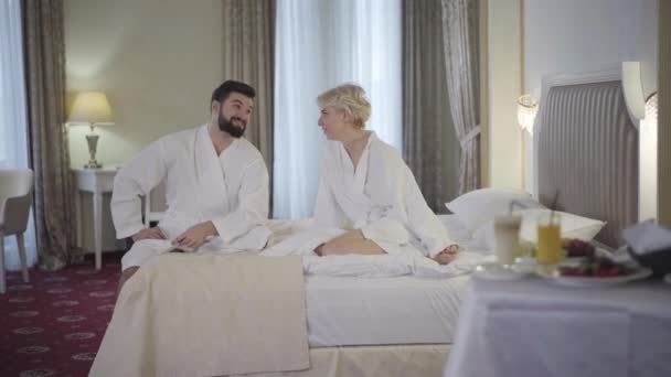 Широкий снимок счастливой белой пары, говорящей на кровати в отеле, как неизвестный официант, катящийся за столом с завтраком. Возбужденные мужчина и женщина получают фрукты по утрам. Обслуживание номеров, туризм, роскошь , — стоковое видео