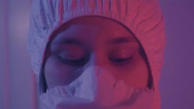 Koruyucu gözlük takan yüz maskeli laboratuvar asistanının aşırı yakın çekimi. Antiviral giysili beyaz kadın portresi laboratuvarda kırmızı ışıkla kameraya bakıyor. Covid-19, salgın hastalık.