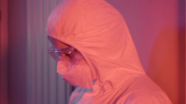 Duvardan aşağı kırmızı ışıkla inen yorgun ve depresif sağlık görevlisinin yan görüntüsü. Koruyucu takım elbiseli ve gözlüklü umutsuz beyaz kadın portresi. Covid-19 salgını, virüs.