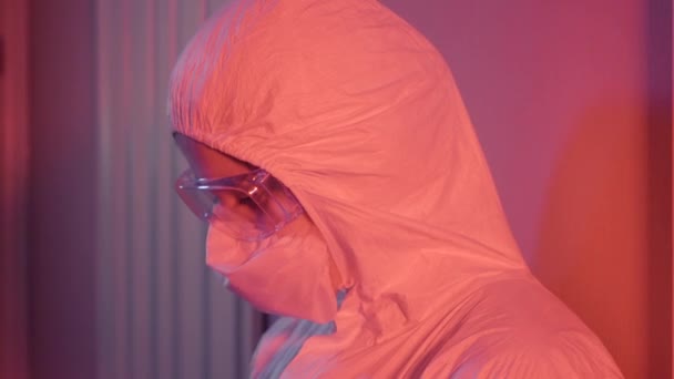 Boční pohled vyčerpaného zdravotníka, který se pohybuje po stěně v místnosti s červeným světlem. Portrét mladé bělošky v ochranném obleku a brýlích. Covid-19 pandemie, virus. — Stock video