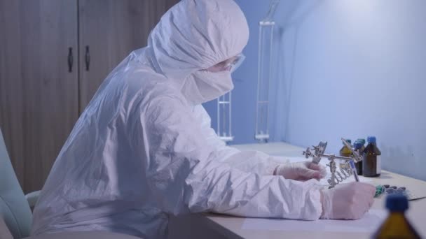 Вид сбоку на уверенного кавказского медработника, осматривающего ватный тампон с помощью лупы. Женщина в белом противовирусном защитном костюме пишет и думает. Пандемия Ковид-19, разработка вакцины . — стоковое видео