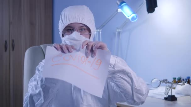 Retrato de doctora caucásica seria en traje protector rasgando hoja de papel con palabra Covid-19. Lucha contra la infección por coronavirus, cuarentena pandémica, bloqueo global . — Vídeo de stock