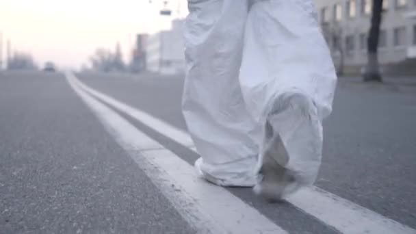 Voeten van viroloog in wit beschermend pak lopen langs lege stadsweg. Camera volgende persoon wandelen in het midden van de snelweg op Covid-19 lockdown. Quarantaine van het coronavirus, pandemie. — Stockvideo