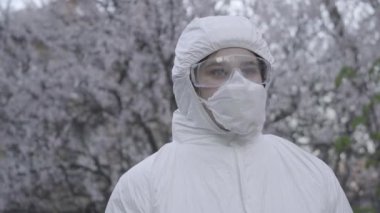 Antiviral takım elbiseli düşünceli beyaz kadın ve çiçek açan ağacın arkasında koruyucu gözlükler. Genç doktor ya da virüs uzmanı. Koronavirüs karantinasında. Covid-19 salgını, tecrit.