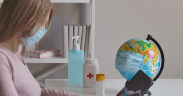 Vista lateral de mujer caucásica confiada en máscara facial sentada en la mesa en casa y encendiendo la tableta. Globo con máscara de pie en el fondo. Bloqueo de Covid-19, trabajo lejano. Sede del cine 4k ProRes . — Vídeos de Stock