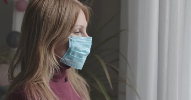 Zijaanzicht close-up van trieste blanke vrouw met gezichtsmasker kijkend uit het raam van Covid-19 lockdown. Portret van een volwassen dame die de lentedag binnen doorbrengt op coronavirus quarantaine. Cinema 4k Protres Hoofdkwartier.. — Stockvideo